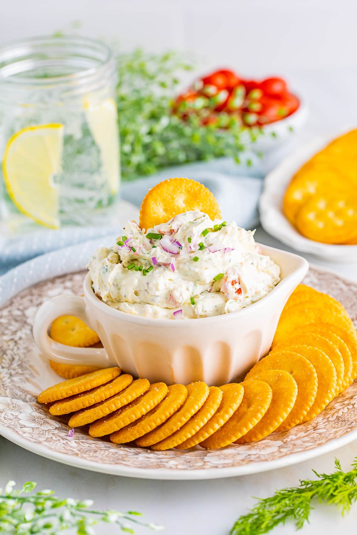 Smoked salmon cream cheese dip served in an ivory measuring cup on a vintage plate with crackers, iced water with lemon, tomatoes, and blue linen in background