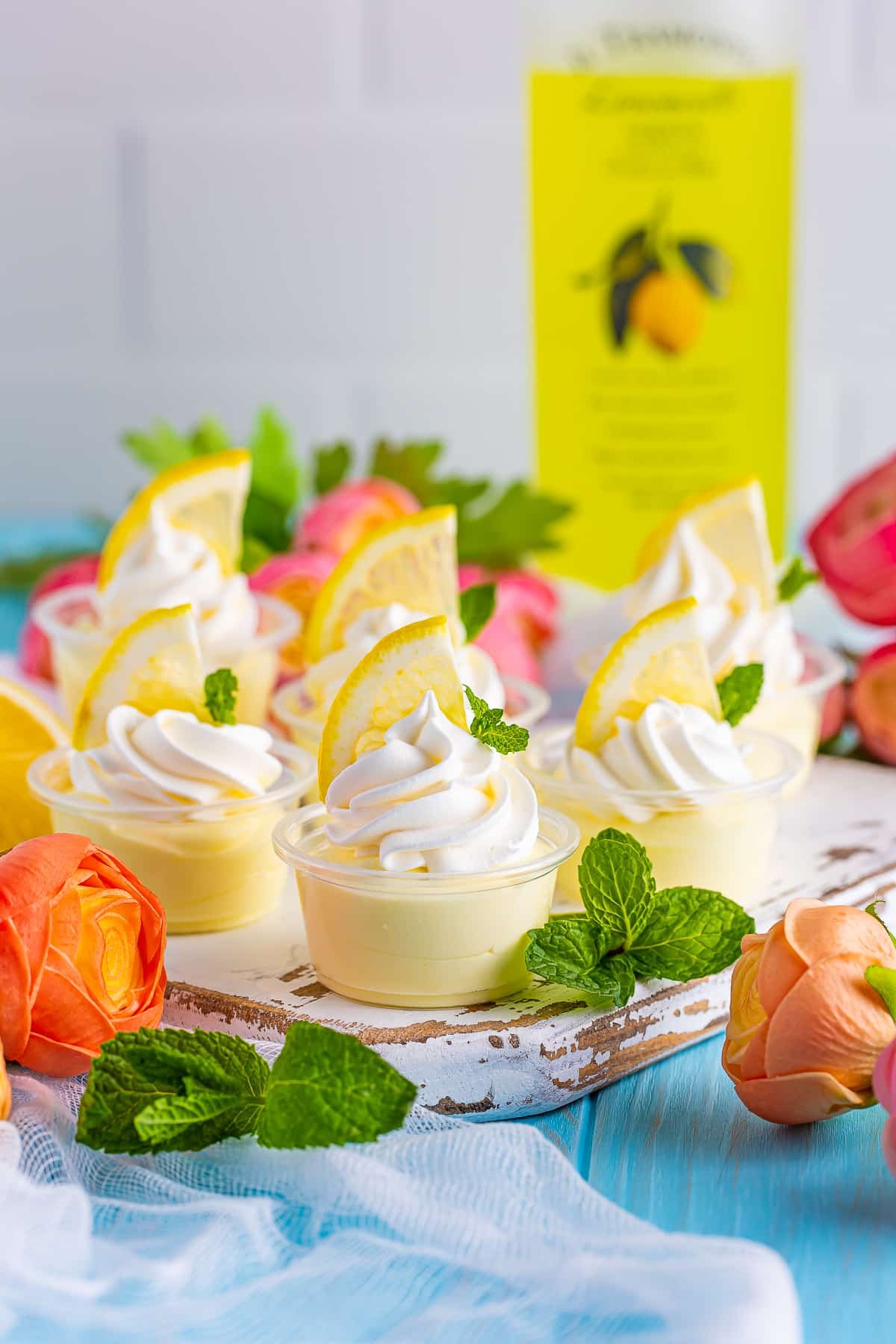 lemon pudding shots on a white wooden serving platter sitting on a blue wood table top, fresh flowers, mint, and lemon