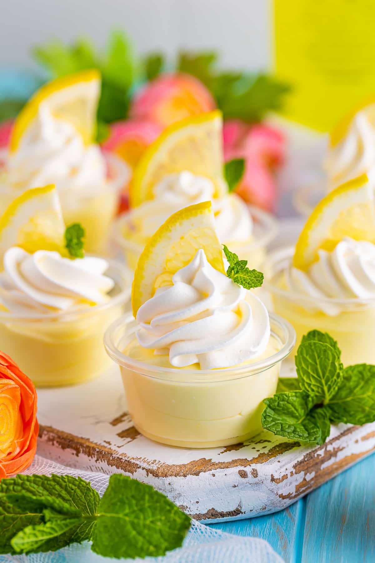 Upclose image of lemon pudding shot on a white wooden serving platter sitting on blue wood table top, fresh mint, lemons, and flowers