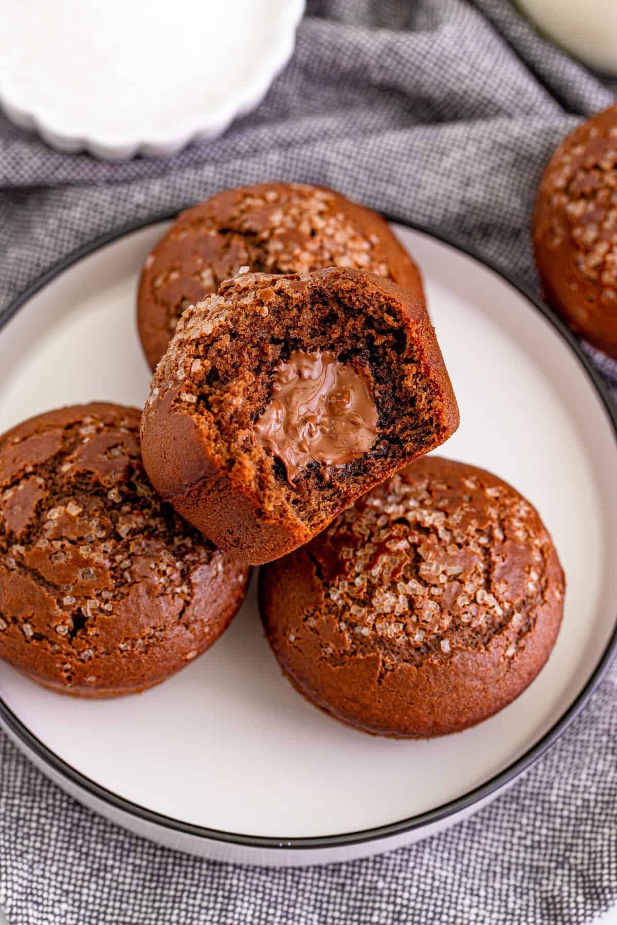 Four nutella muffins on a white plate with black rim with one on top with a bite taken out with grey kitchen towel