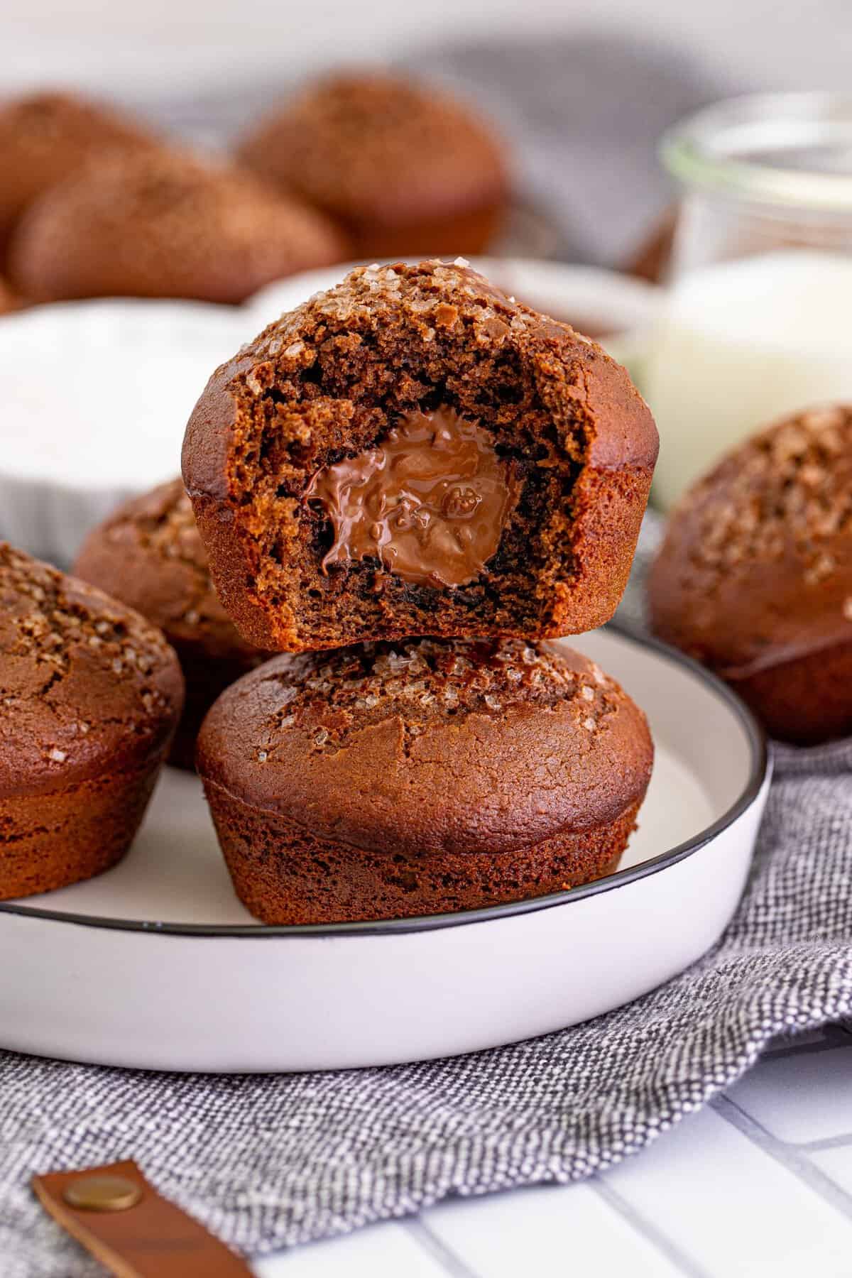 Two nutella muffins stacked on a white plate with black rim sitting on a grey kitchen towel, the muffin on top has a bite taken out so you can see the nutella filling