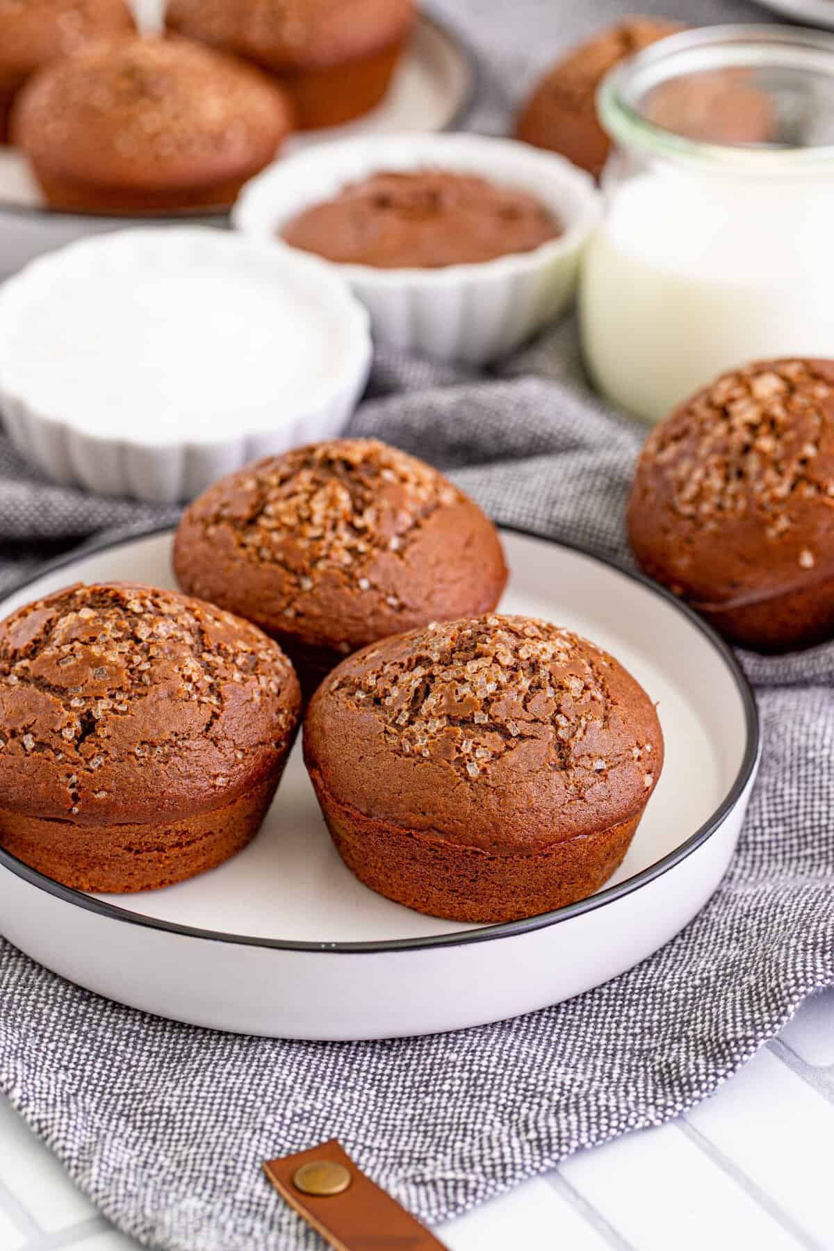 Nutella muffins sitting on a white plate with black rim on a grey kitchen towel, glass of milk and nutella in the background
