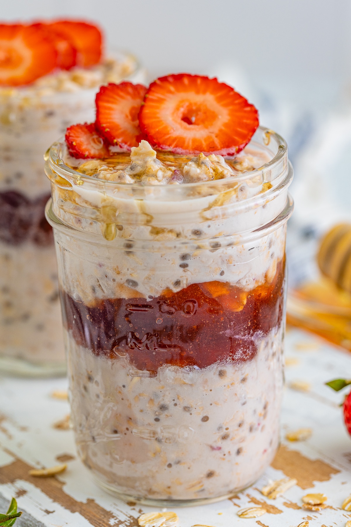 Strawberry Overnight oats shown layered in a mason jar on a white wooden serving platter, striped blue linen, and honey dipper in the background
