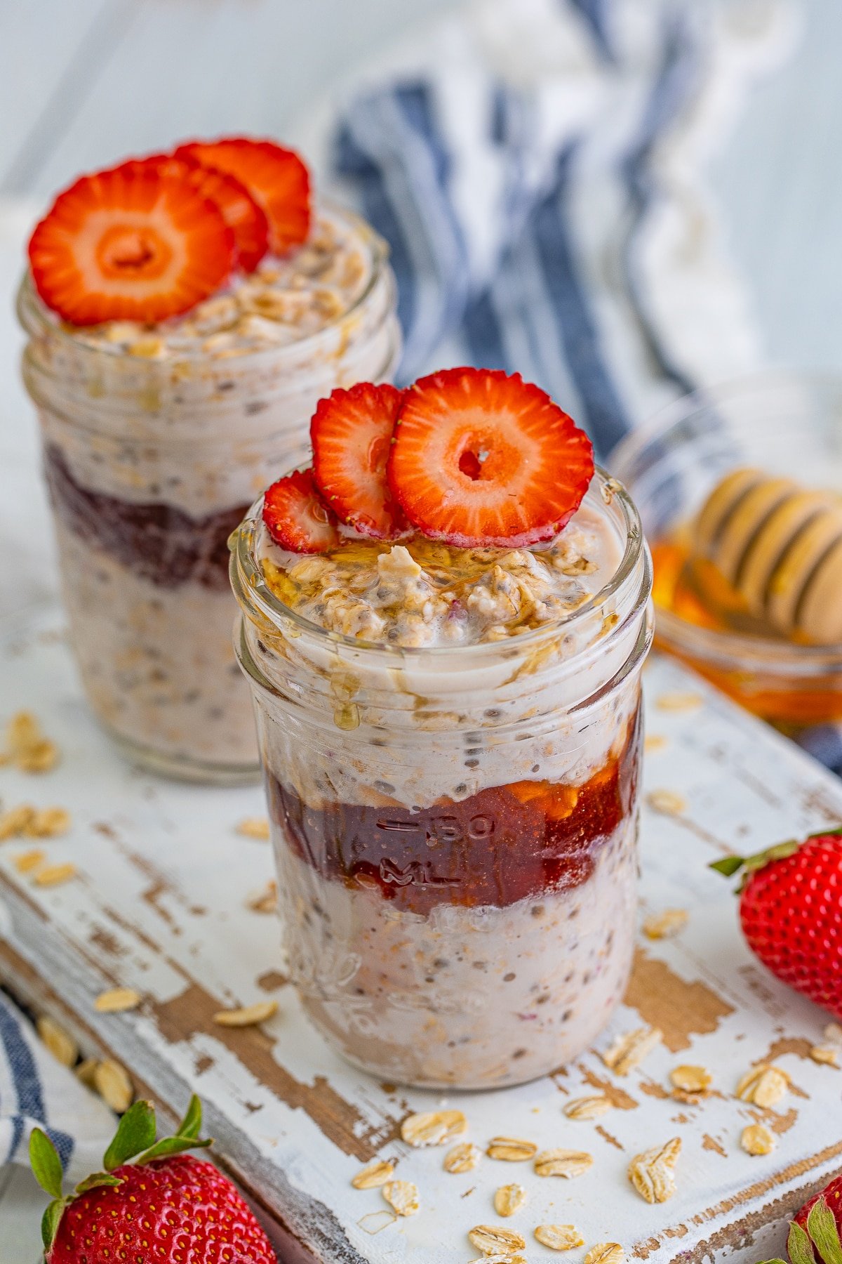 Strawberry Overnight oats shown layered in a mason jar on a white wooden serving platter, fresh strawberries, striped blue linen, and honey dipper in the background