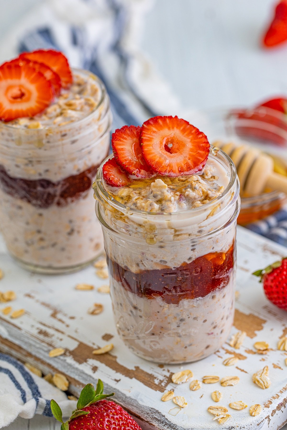Strawberry Overnight oats shown layered in a mason jar on a white wooden serving platter, fresh strawberries, striped blue linen, and honey dipper in the background