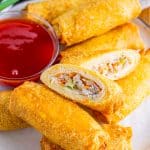 Crispy air fryer egg rolls on a plate, one cut open to show the filling, with sweet chili dipping sauce.