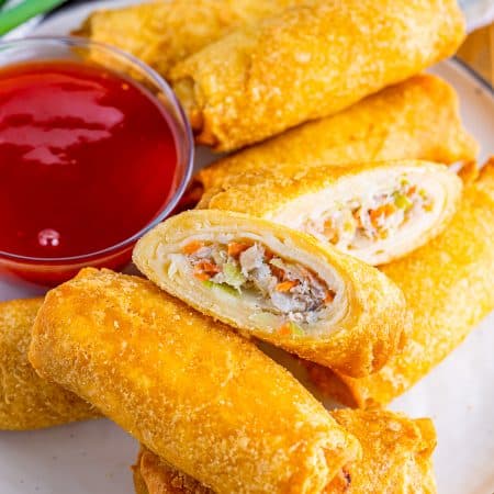 Crispy air fryer egg rolls on a plate, one cut open to show the filling, with sweet chili dipping sauce.