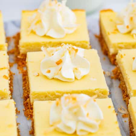 Coconut cheesecake bars topped with whipped cream swirls and toasted coconut arranged on parchment paper.