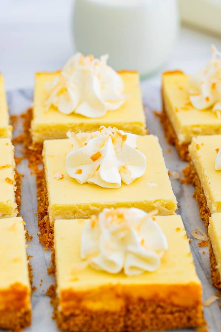 Coconut cheesecake bars topped with whipped cream swirls and toasted coconut arranged on parchment paper.