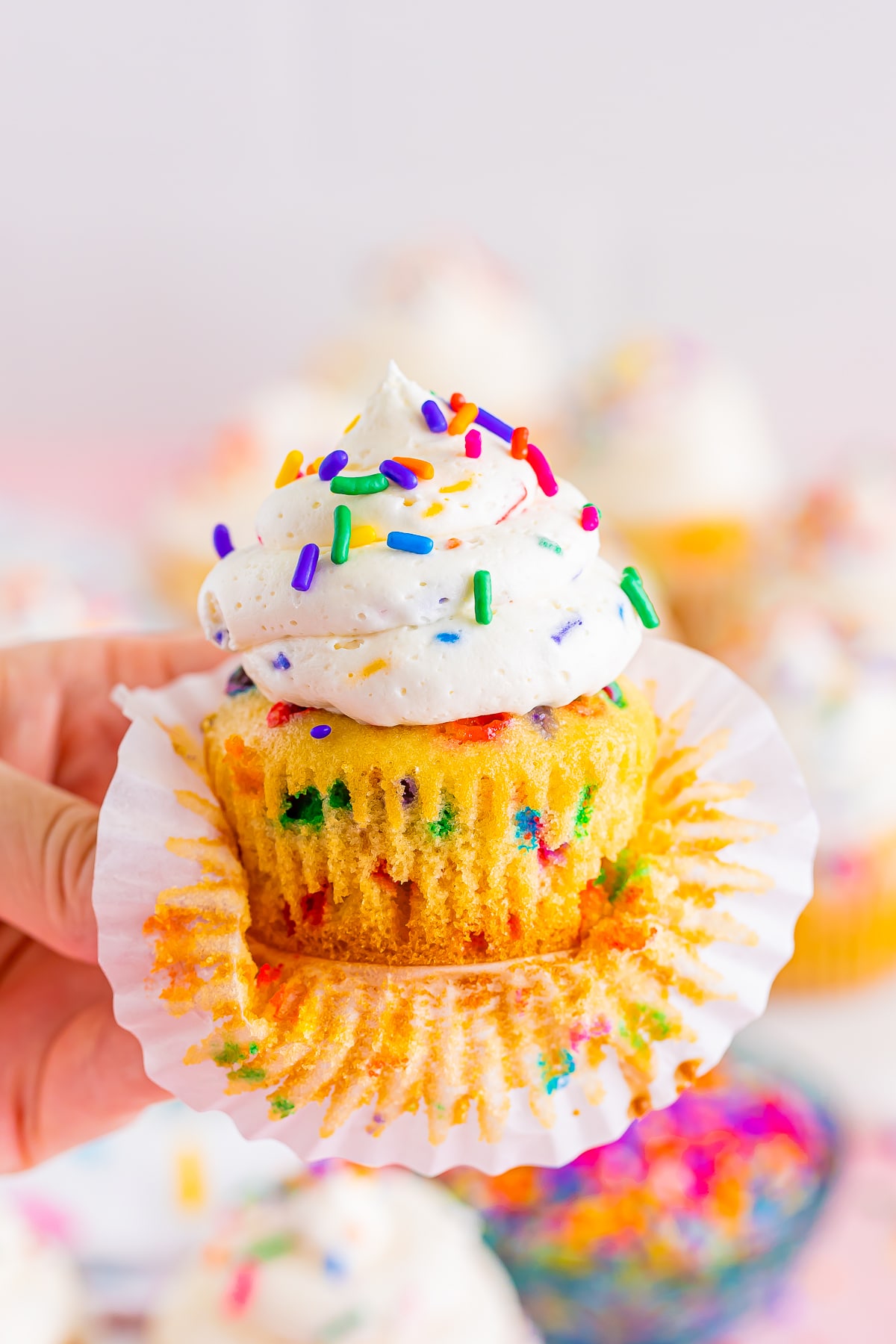 A hand holding a funfetti cupcake in air with the paper liner coming off