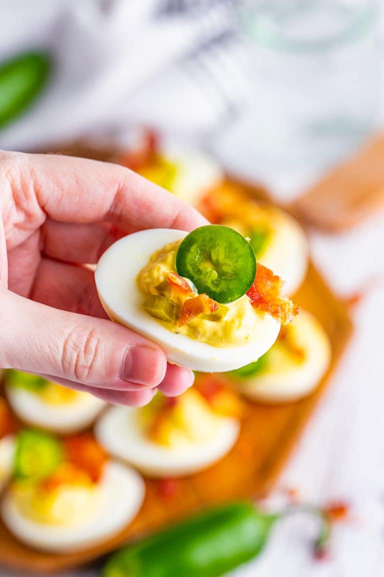 A wood board of jalapeno deviled eggs topped with crispy bacon and fresh jalapeno slices.