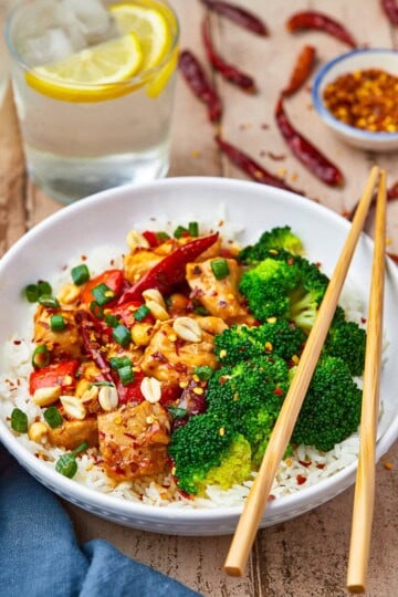 A bowl of kung pao chicken with bell peppers and peanuts served over rice with broccoli.