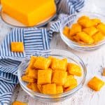 A glass bowl of homemade Cheez-Its with a block of cheddar cheese.