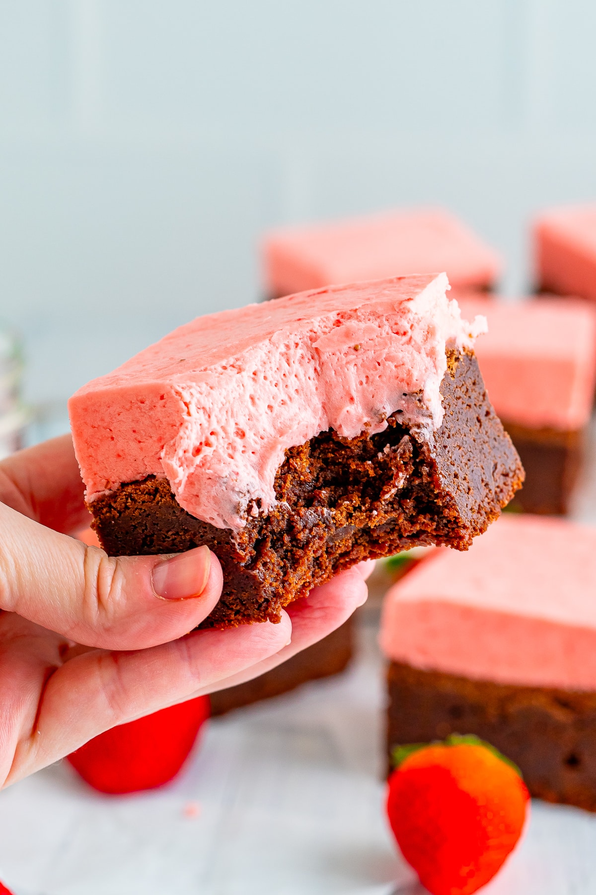 A HAND HOLDING A BROWNIE WITH STRAWBERRY FROSTING IN AIR WITH A BITE TAKEN OUT