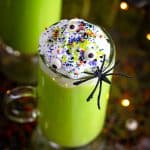 A tall glass of green Witches Brew hot chocolate topped with whipped cream, candy eyes, sprinkles, and a plastic spider in front of a slow cooker.
