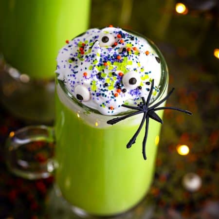 A tall glass of green Witches Brew hot chocolate topped with whipped cream, candy eyes, sprinkles, and a plastic spider in front of a slow cooker.