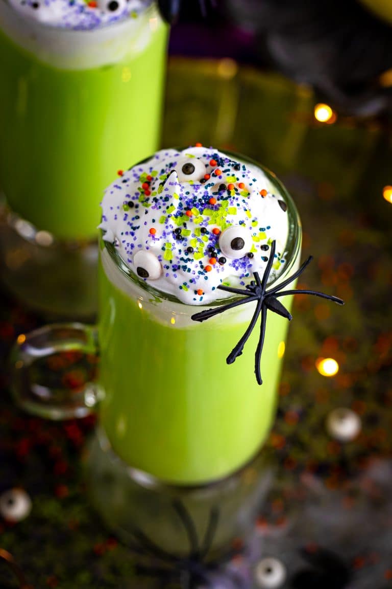 A tall glass of green Witches Brew hot chocolate topped with whipped cream, candy eyes, sprinkles, and a plastic spider in front of a slow cooker.