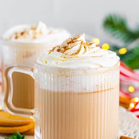 A Gingerbread Latte in a glass mug topped with whipped cream and cinnamon next to gingersnap cookies.
