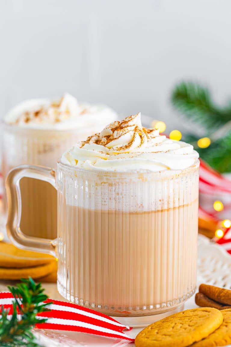 A Gingerbread Latte in a glass mug topped with whipped cream and cinnamon next to gingersnap cookies.