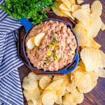 A skillet of creamy dill pickle cheeseburger dip garnished with parsley and pickles, surrounded by ridged potato chips.