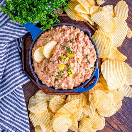 A skillet of creamy dill pickle cheeseburger dip garnished with parsley and pickles, surrounded by ridged potato chips.