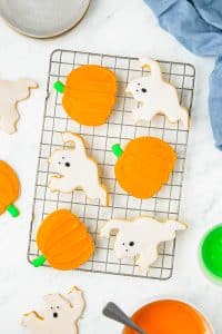 Decorated Halloween sugar cookies shaped like orange pumpkins and white ghosts cooling on a wire rack.