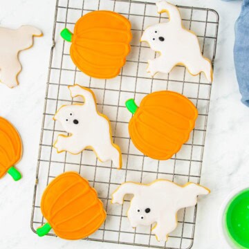 Decorated Halloween sugar cookies shaped like orange pumpkins and white ghosts cooling on a wire rack.