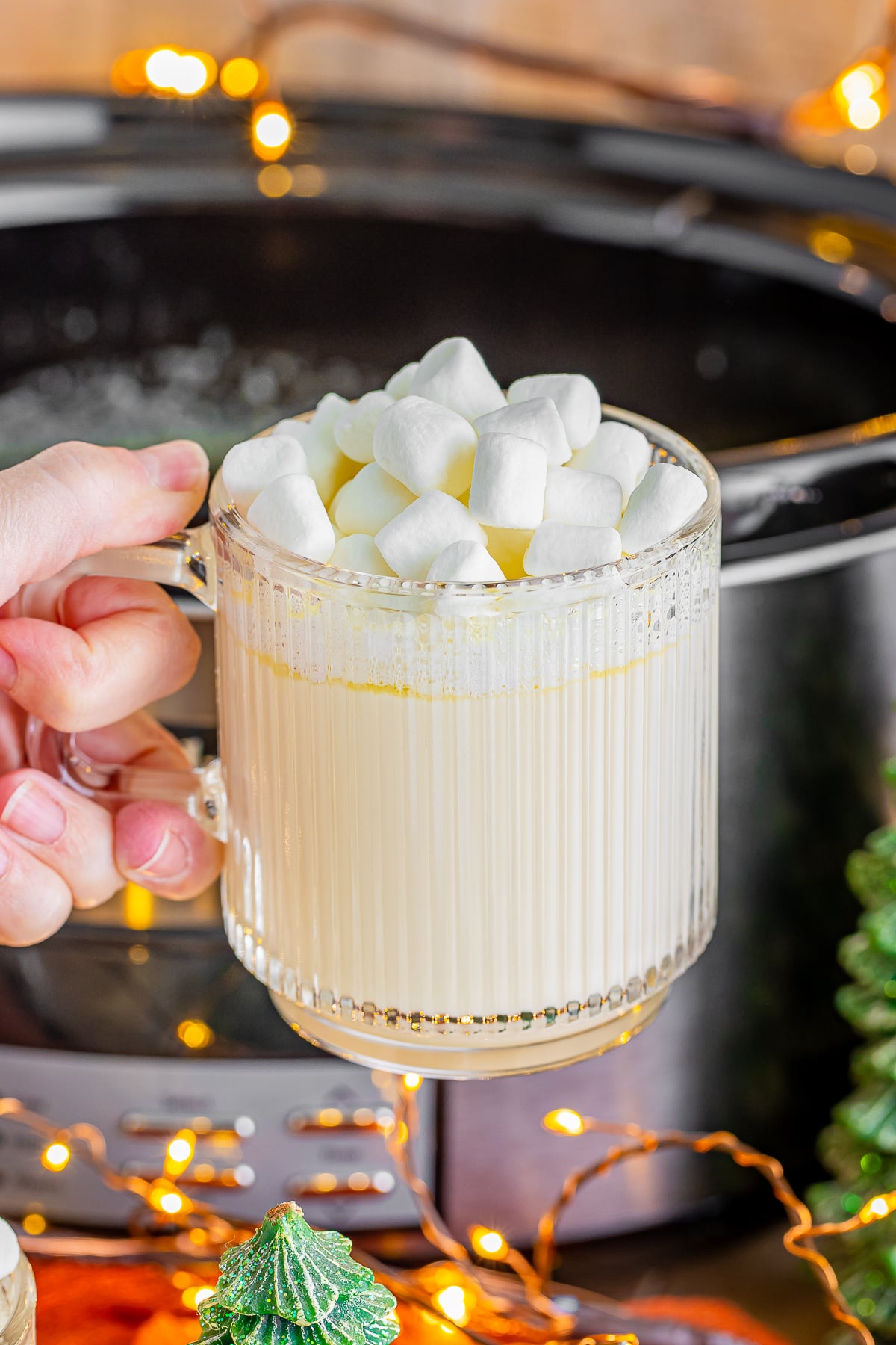 a hand holding up white hot chocolate in front of a slow cooker