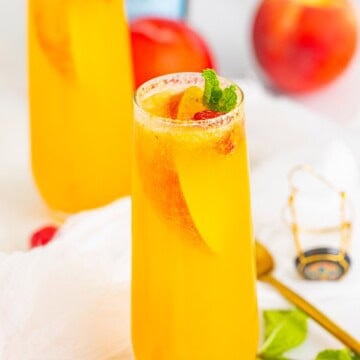 Two peach bellini cocktails in champagne flutes garnished with peach slices, raspberries, and mint.