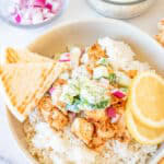 A Greek Chicken Bowl with rice, seared chicken, tzatziki, red onion, and lemon slices with pita bread on the side.