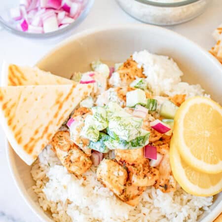 A Greek Chicken Bowl with rice, seared chicken, tzatziki, red onion, and lemon slices with pita bread on the side.