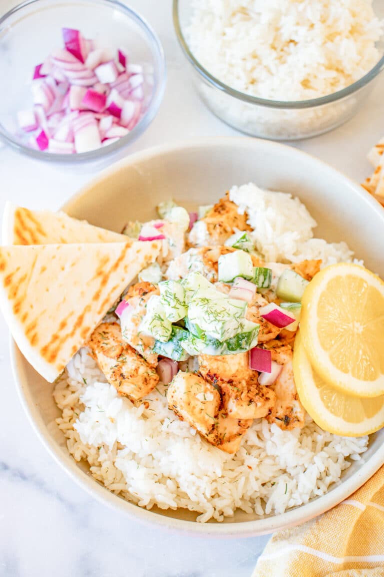 A Greek Chicken Bowl with rice, seared chicken, tzatziki, red onion, and lemon slices with pita bread on the side.