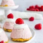A mini lemon cheesecake topped with whipped topping and a fresh raspberry on a white plate.