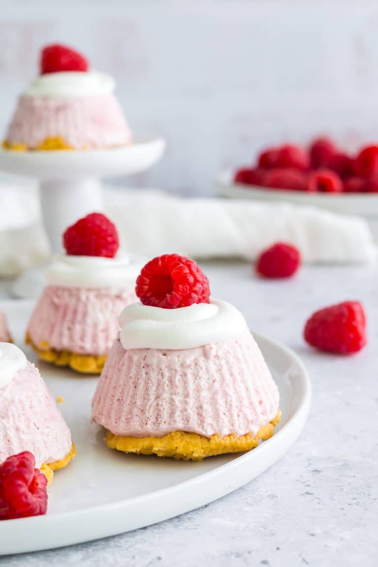 A mini lemon cheesecake topped with whipped topping and a fresh raspberry on a white plate.