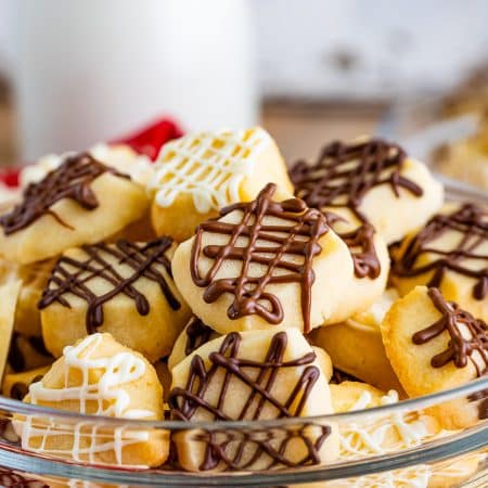 A glass bowl piled high with Christmas shortbread cookies drizzled in dark, milk, and white chocolate next to a bottle of milk.