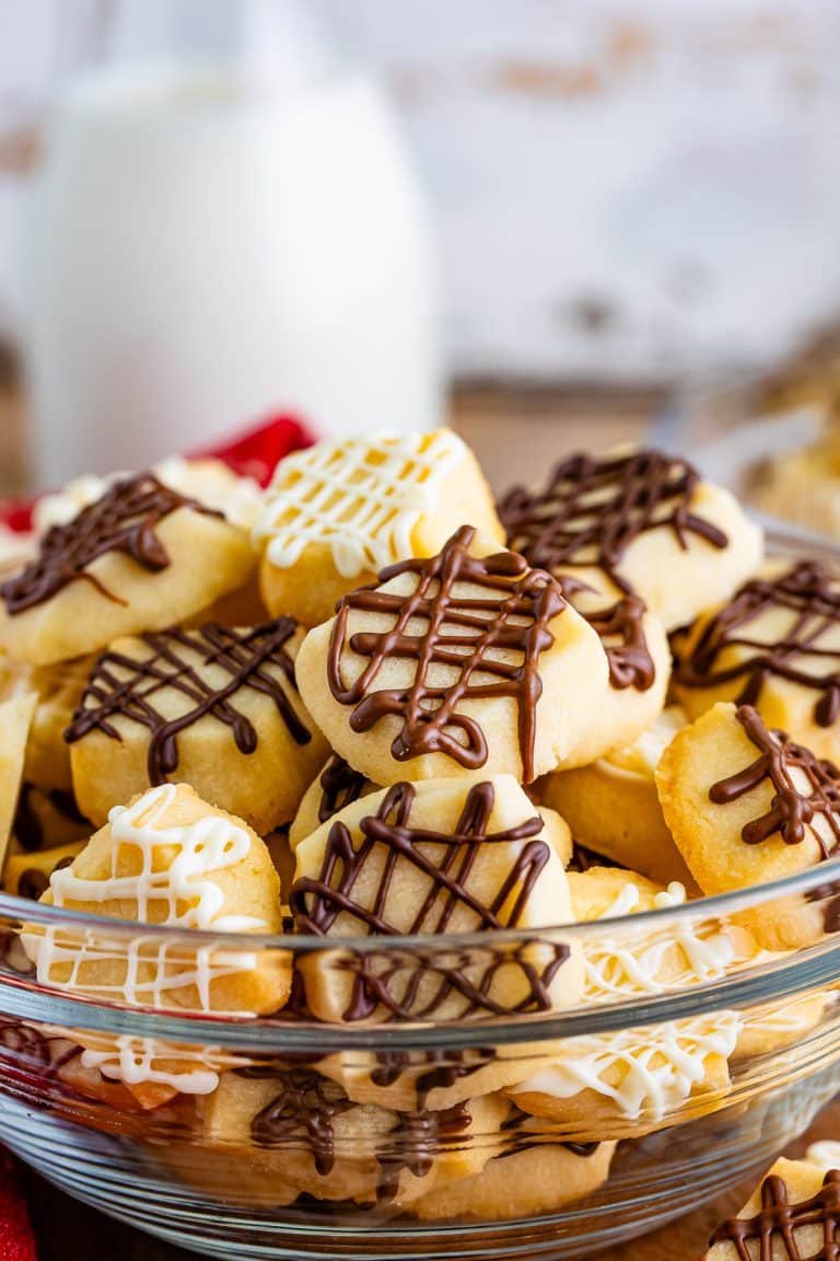 A glass bowl piled high with Christmas shortbread cookies drizzled in dark, milk, and white chocolate next to a bottle of milk.