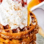 Maple syrup being poured over a stack of Gingerbread Waffles topped with whipped cream and cinnamon.