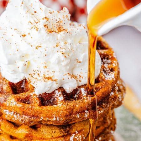 Maple syrup being poured over a stack of Gingerbread Waffles topped with whipped cream and cinnamon.