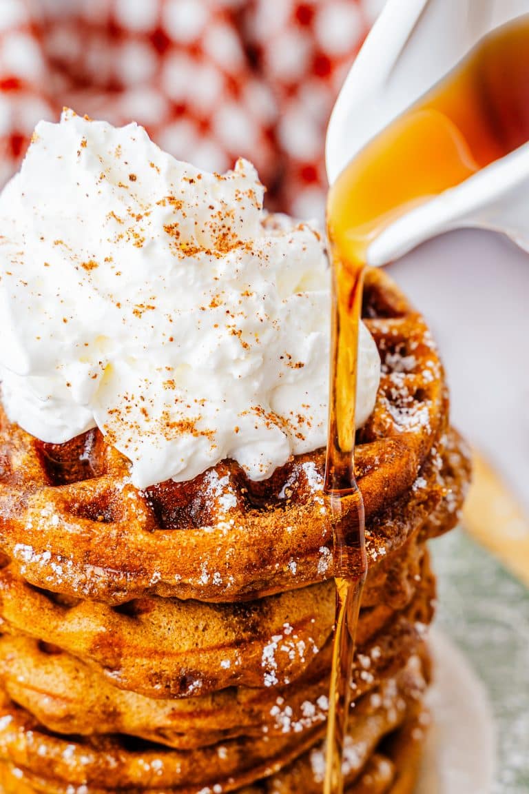 Maple syrup being poured over a stack of Gingerbread Waffles topped with whipped cream and cinnamon.