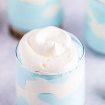 A glass of blue and white jello cloud mousse swirled together like a cloudy sky, topped with whipped cream.
