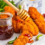 Crispy Hot Honey Chicken tenders piled next to a jar of hot honey with a wooden dipper and fresh chiles.
