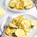 A baking dish of cheesy scalloped zucchini with a wooden spoon scooping a creamy, cheesy serving.
