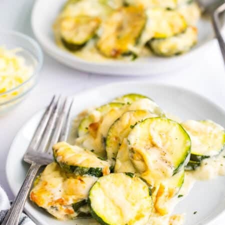 A baking dish of cheesy scalloped zucchini with a wooden spoon scooping a creamy, cheesy serving.