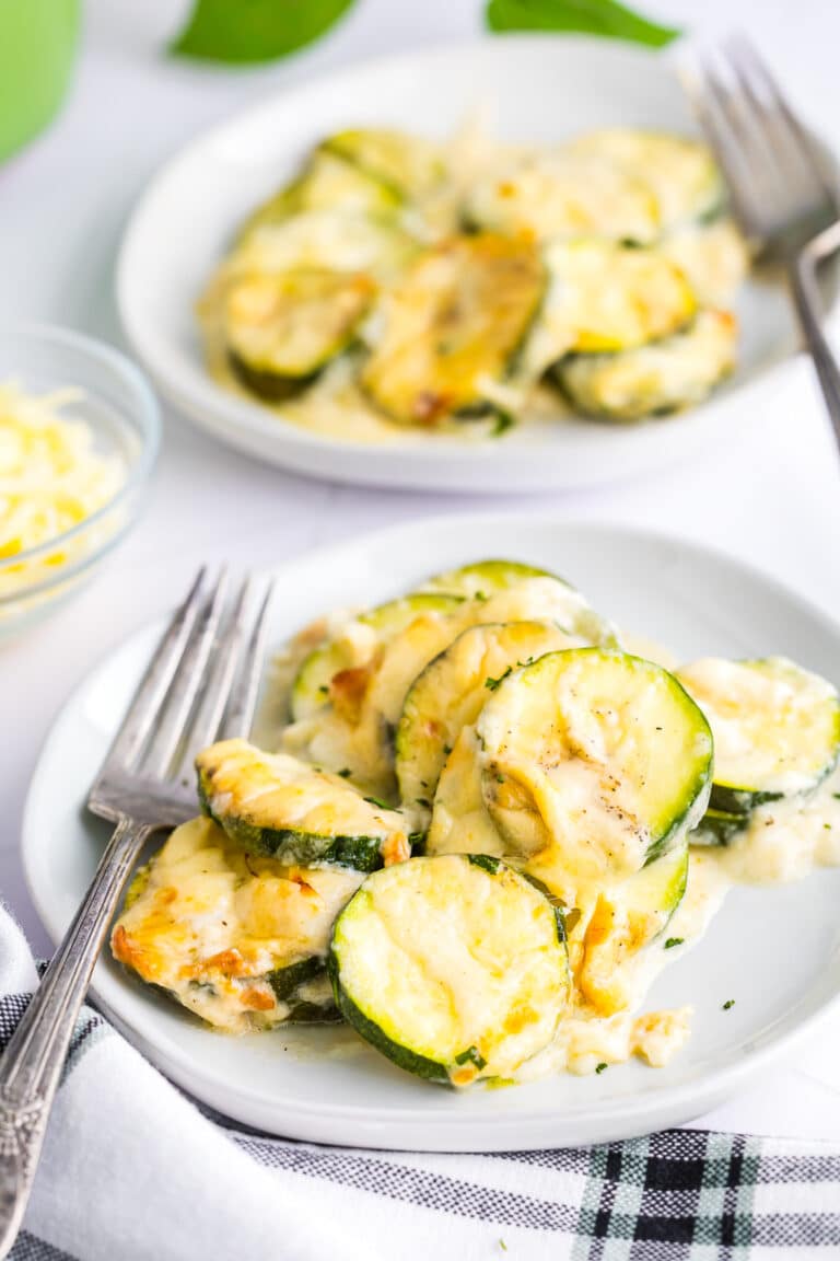 A baking dish of cheesy scalloped zucchini with a wooden spoon scooping a creamy, cheesy serving.