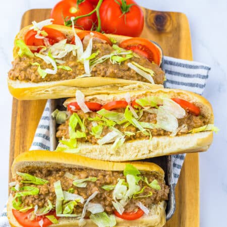 Three slow cooker chopped cheese sandwiches on a wooden board topped with lettuce and tomato.