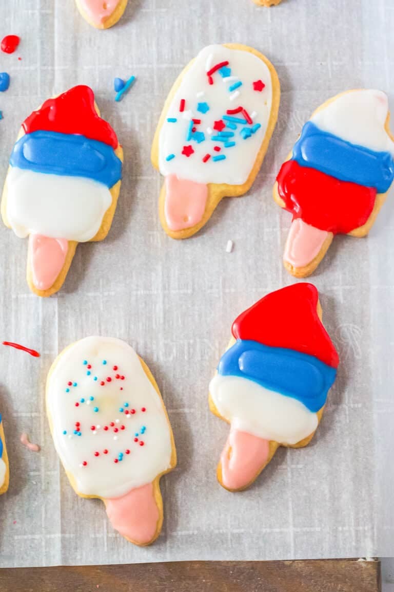 Popsicle shaped cut out sugar cookies decorated with red, white, and blue icing on parchment.