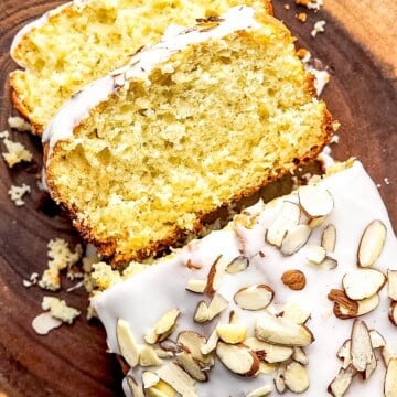 Sliced glazed almond cake topped with sliced almonds on a wood board.