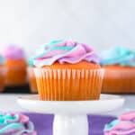 A cherry cupcake with a swirled pink, purple, and turquoise tie dye frosting on a small white cake stand.