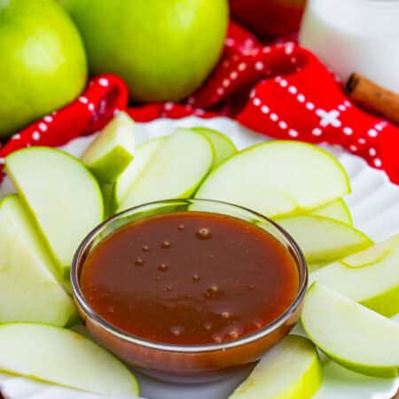 Caramel sauce in a glass bowl surrounded by green apple slices on a white plate.