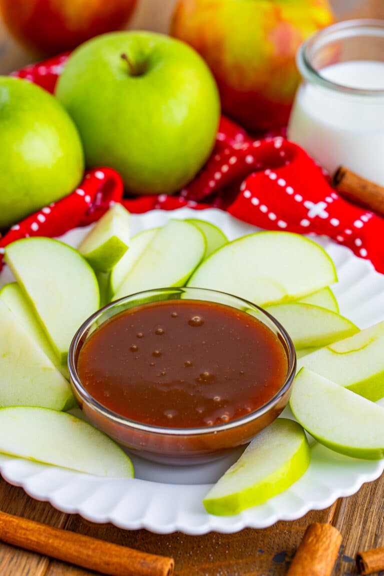 Caramel sauce in a glass bowl surrounded by green apple slices on a white plate.